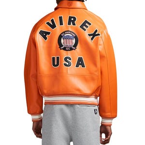 May include: An orange leather bomber jacket featuring the text AVIREX in black letters arched across the upper back, a circular patch with a US flag design, and the text USA below. The jacket has a ribbed collar and cuffs with white stripes.