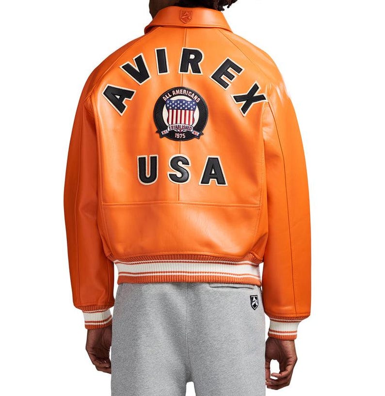 May include: An orange leather bomber jacket featuring the text AVIREX in black letters arched across the upper back, a circular patch with a US flag design, and the text USA below. The jacket has a ribbed collar and cuffs with white stripes.