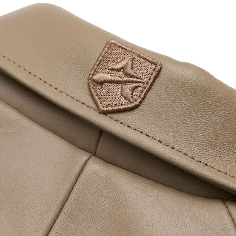 May include: A light brown leather item featuring a smooth, matte texture and visible stitching along the edges. Centered on the upper flap is a small, shield-shaped patch with an embroidered emblem in a matching brown thread color.