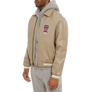 May include: A beige leather jacket with a collar, a full-length front zipper, and ribbed cuffs. The chest features a patch with the text AVIREX and an American flag graphic. The jacket is worn over a grey hooded sweatshirt and grey sweatpants.