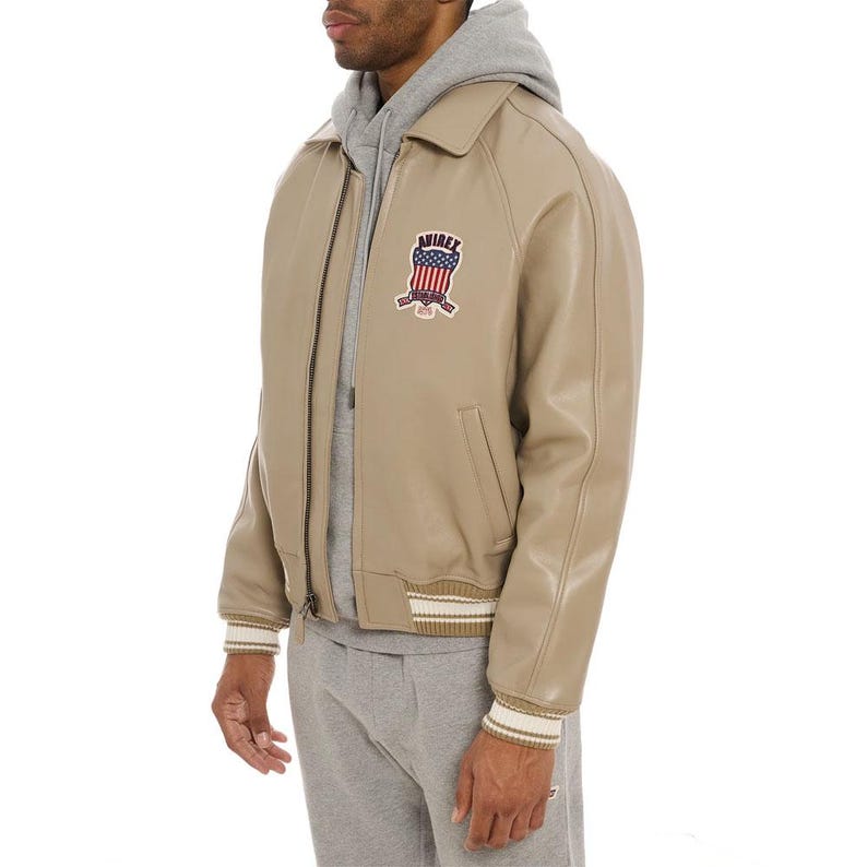 May include: A beige leather jacket with a collar, a full-length front zipper, and ribbed cuffs. The chest features a patch with the text AVIREX and an American flag graphic. The jacket is worn over a grey hooded sweatshirt and grey sweatpants.