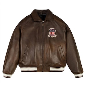 May include: A dark brown leather bomber jacket featuring a front zipper closure, a fold-down collar, and two side pockets. A patch on the left chest displays the text AVIREX above an American flag graphic with the text ESTABLISHED 1975 below.