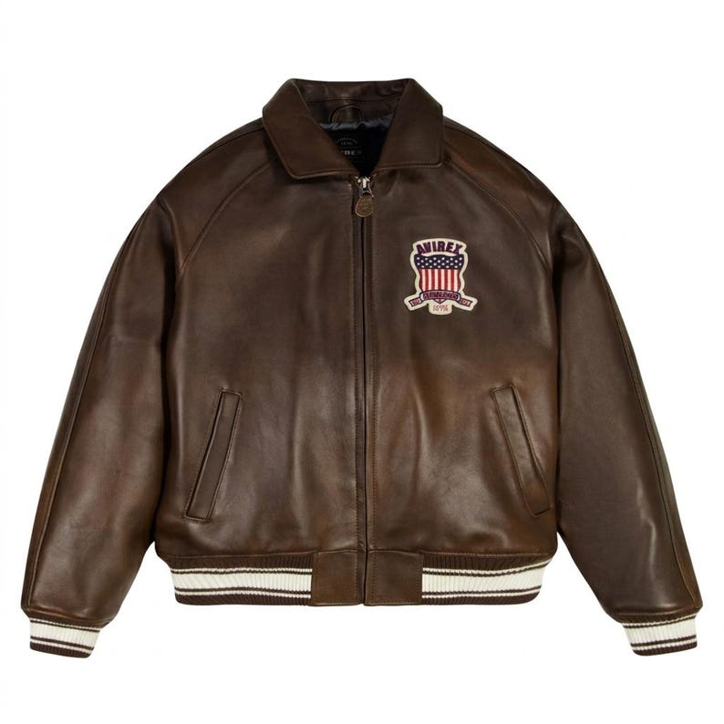 May include: A dark brown leather bomber jacket featuring a front zipper closure, a fold-down collar, and two side pockets. A patch on the left chest displays the text AVIREX above an American flag graphic with the text ESTABLISHED 1975 below.