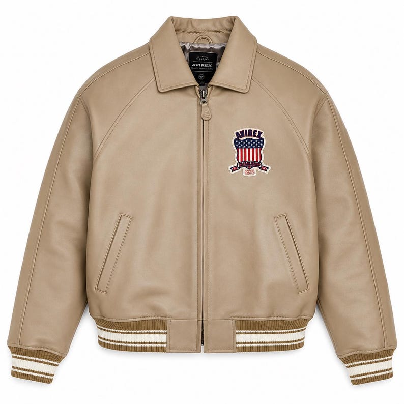May include: A tan leather bomber jacket with a front zipper closure, a pointed collar, and ribbed cuffs and hem featuring white stripes. A patch on the left chest displays the text AVIREX above a shield with red, white, and blue flag details.