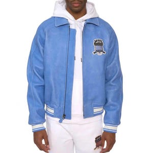 May include: A light blue suede jacket featuring a white collar, a full-length front zipper, and ribbed cuffs with white stripes. An embroidered patch on the left chest displays the text AVIREX ESTABLISHED 1975 above a shield with a flag design.