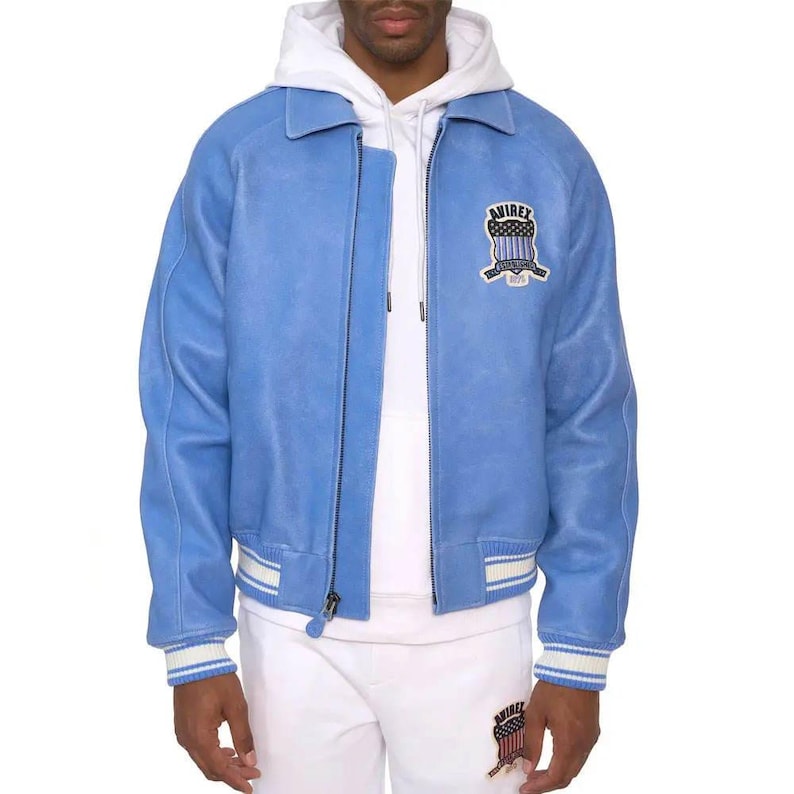 May include: A light blue suede jacket featuring a white collar, a full-length front zipper, and ribbed cuffs with white stripes. An embroidered patch on the left chest displays the text AVIREX ESTABLISHED 1975 above a shield with a flag design.