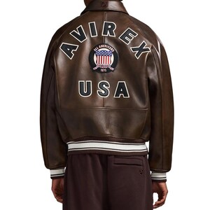 May include: A brown leather jacket featuring the text AVIREX in an arc above a circular patch with an American flag and the text ALL AMERICANS ESTABLISHED 1975. The word USA is printed in large letters below the patch. The jacket has a ribbed hem.