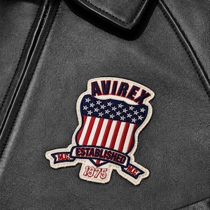 May include: A black leather jacket featuring an embroidered patch on the chest. The patch displays a shield with red and white stripes and white stars on a blue background, with the text AVIREX ESTABLISHED 1975 and M.C. in red and blue.