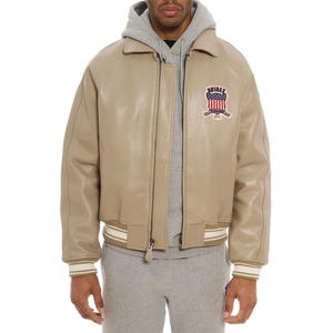 May include: A beige leather bomber jacket featuring a collared neckline, a front zipper closure, and striped ribbed cuffs and hem. A patch on the left chest displays the text AVIREX above a graphic of the American flag and the words ESTABLISHED 1975.