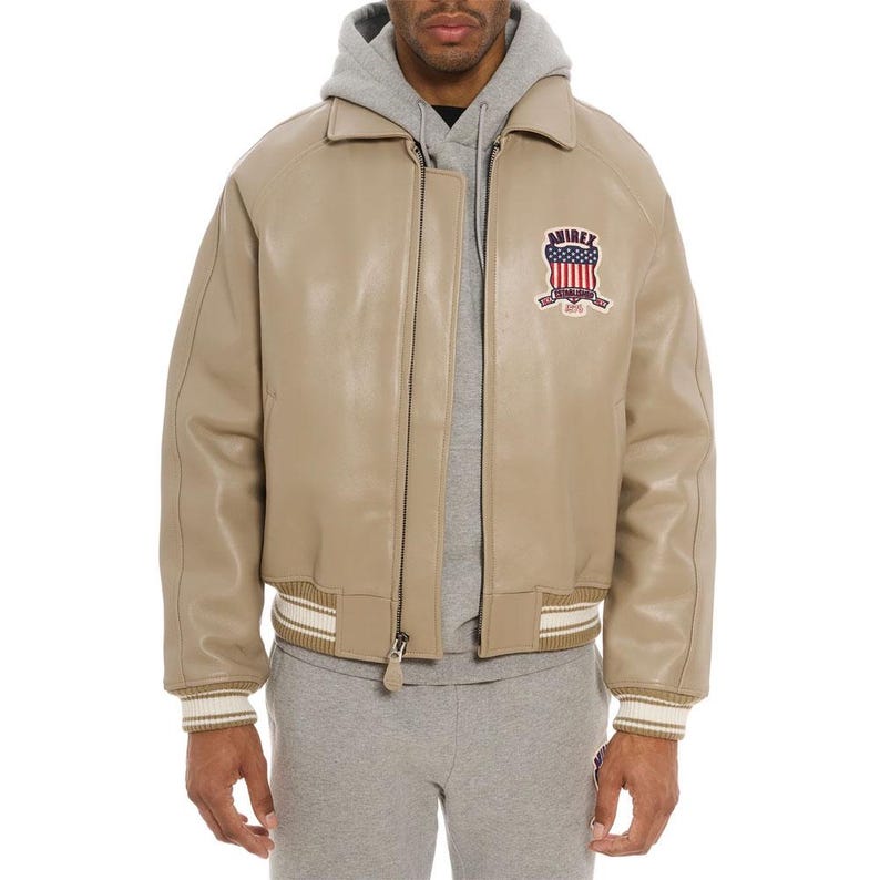 May include: A beige leather bomber jacket featuring a collared neckline, a front zipper closure, and striped ribbed cuffs and hem. A patch on the left chest displays the text AVIREX above a graphic of the American flag and the words ESTABLISHED 1975.