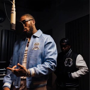 May include: A light blue leather bomber jacket with a white and blue Avirex patch on the chest, featuring a classic collar, ribbed cuffs with white and blue stripes, and a front zipper closure, worn by a person in a recording studio setting.