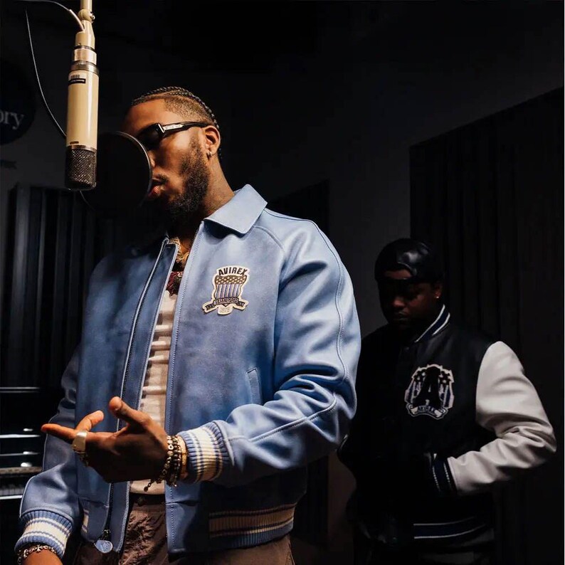 May include: A light blue leather bomber jacket with a white and blue Avirex patch on the chest, featuring a classic collar, ribbed cuffs with white and blue stripes, and a front zipper closure, worn by a person in a recording studio setting.