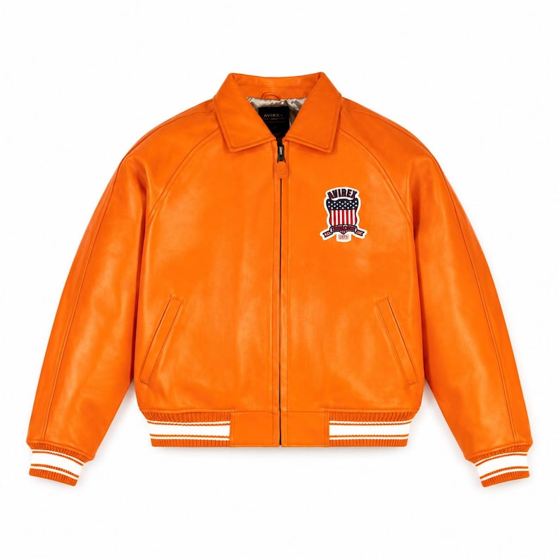 May include: A bright orange leather jacket featuring a full-length front zipper, a classic collar, and ribbed cuffs and hem with white stripes. On the left chest, there is a patch displaying the text AVIREX with a shield and American flag.