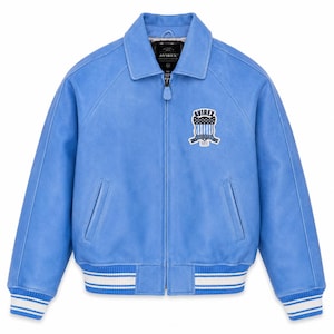 May include: A light blue jacket with a full-length front zipper, a classic collar, and ribbed cuffs and hem featuring white stripes. The chest displays a patch with the text AVIREX and a shield graphic. The interior label also reads AVIREX.