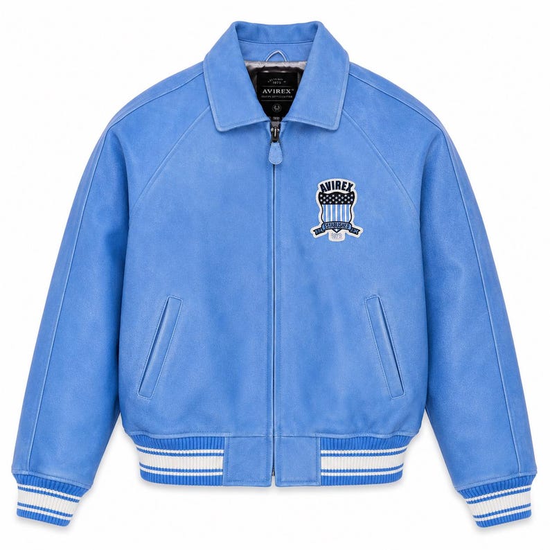 May include: A light blue jacket with a full-length front zipper, a classic collar, and ribbed cuffs and hem featuring white stripes. The chest displays a patch with the text AVIREX and a shield graphic. The interior label also reads AVIREX.