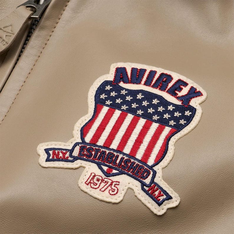 May include: A tan leather jacket featuring an embroidered patch with the text AVIREX, ESTABLISHED, 1975, and N.Y. The patch displays a shield design with a blue field of white stars above red and white vertical stripes, all outlined in cream thread.