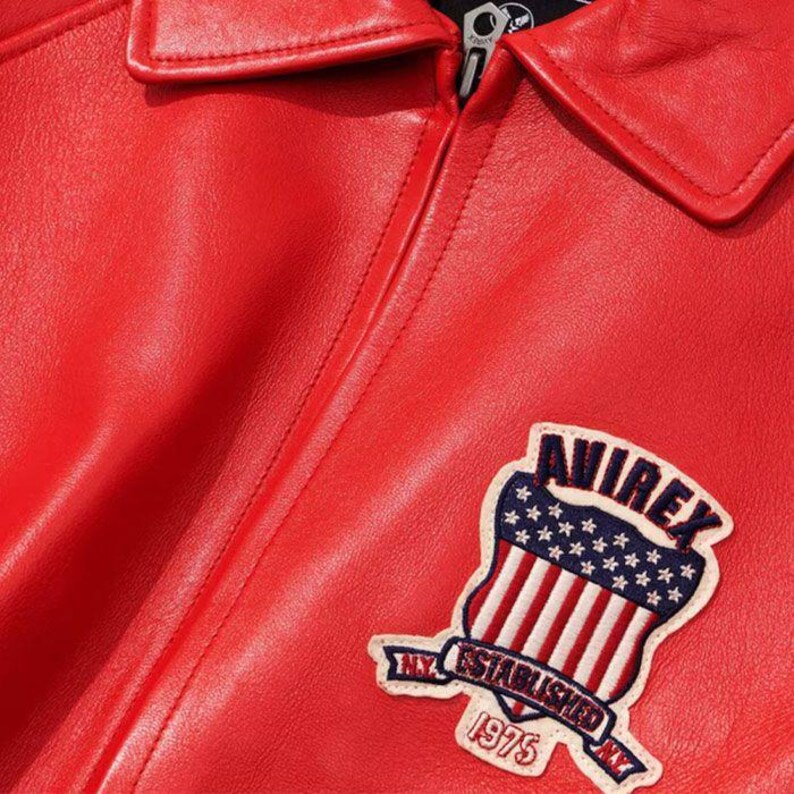 Puede incluir: Una chaqueta de cuero rojo brillante con cremallera frontal completa y cuello en punta. Un parche bordado en el pecho muestra un escudo con estrellas y rayas, el texto AVIREX, ESTABLISHED 1975 y NY sobre un fondo de color crema.
