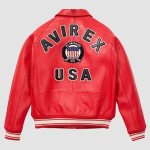 Puede incluir: Una chaqueta de cuero rojo brillante con el texto AVIREX en letras negras arqueadas en la parte superior de la espalda. Debajo del texto hay un parche circular con una bandera de EE. UU. y el texto ALL AMERICANS ESTABLISHED 1975. La palabra USA est&aacute; debajo.