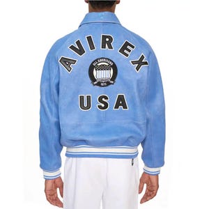 May include: A light blue jacket featuring the text AVIREX in black letters with white outlines arched across the back. Below is a circular patch with the text ALL AMERICANS ESTABLISHED 1975 and the word USA in large black letters with white outlines.