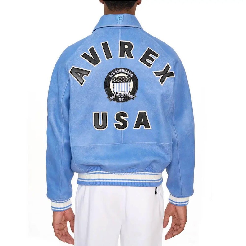 May include: A light blue jacket featuring the text AVIREX in black letters with white outlines arched across the back. Below is a circular patch with the text ALL AMERICANS ESTABLISHED 1975 and the word USA in large black letters with white outlines.