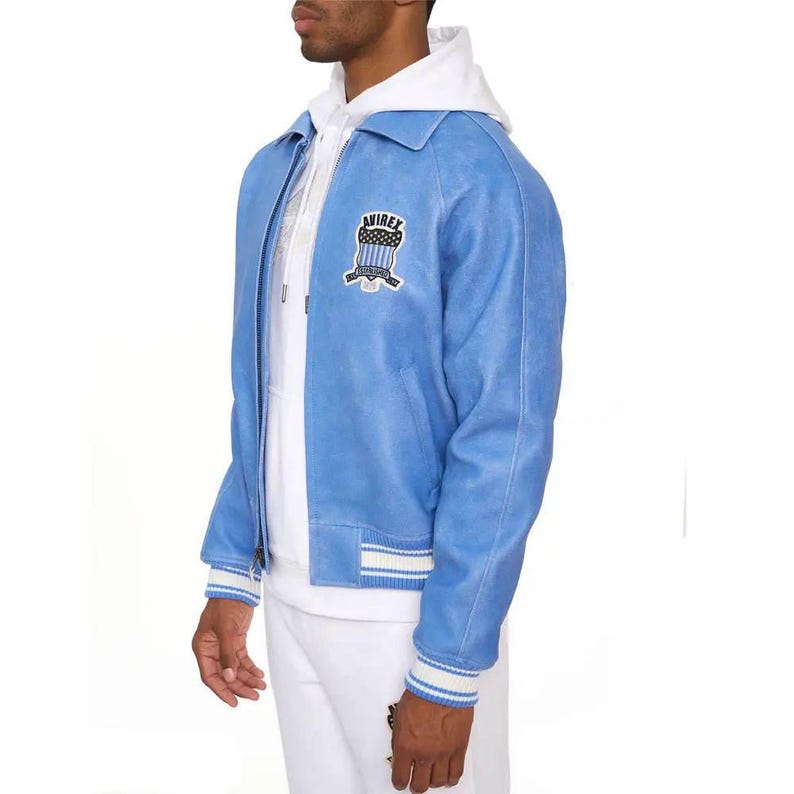 May include: A light blue jacket with a white hoodie underneath. The jacket features a front zipper, a collar, and a patch on the chest with the text AVIREX. The cuffs and hem have white and blue striped ribbed detailing. The jacket is worn over white pants.