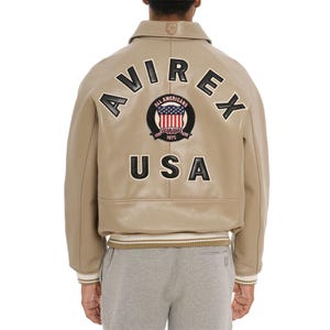 May include: A tan leather bomber jacket featuring the text AVIREX and USA in black lettering on the back. A circular patch in the center displays the American flag with the text ALL AMERICANS ESTABLISHED 1975. The jacket has a ribbed hem and cuffs.