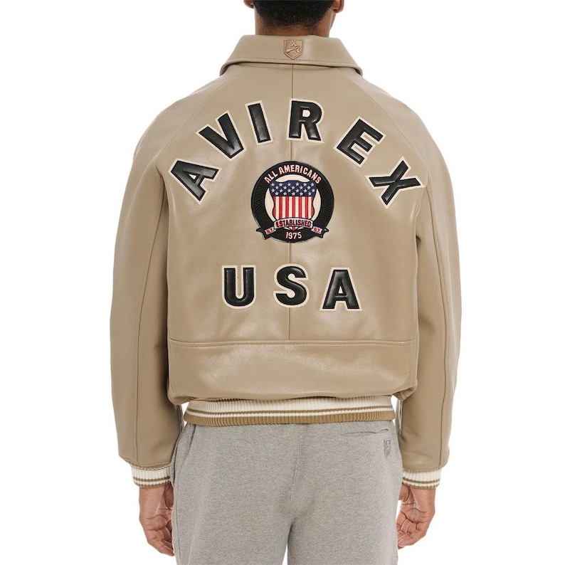 May include: A tan leather bomber jacket featuring the text AVIREX and USA in black lettering on the back. A circular patch in the center displays the American flag with the text ALL AMERICANS ESTABLISHED 1975. The jacket has a ribbed hem and cuffs.