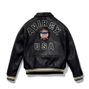 May include: A black leather jacket featuring a white arched AVIREX logo and the text USA on the back. A circular patch with an American flag and the text ALL AMERICANS ESTABLISHED 1875 is centered. The jacket has ribbed cuffs and hem with white stripes.
