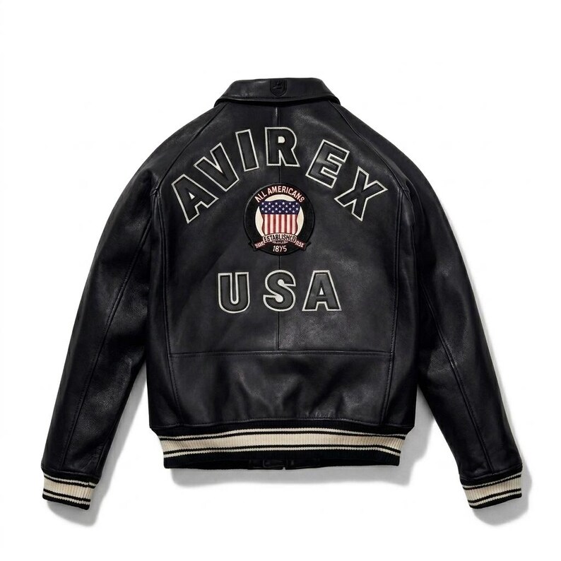 May include: A black leather jacket featuring a white arched AVIREX logo and the text USA on the back. A circular patch with an American flag and the text ALL AMERICANS ESTABLISHED 1875 is centered. The jacket has ribbed cuffs and hem with white stripes.