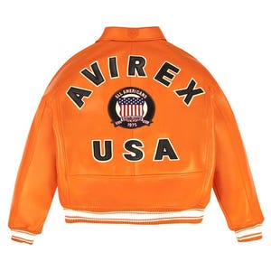 May include: An orange leather bomber jacket featuring the text AVIREX in a black arched font across the upper back. Below this, a circular patch displays the text ALL AMERICANS ESTABLISHED 1975 and a US flag. The word USA appears in black below.