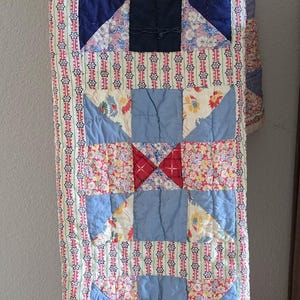 May include: A rectangular patchwork quilt featuring a repeating star pattern composed of various fabric squares. The design includes floral prints, solid navy and light blue blocks, and a vertical border strip with a red and blue star motif.