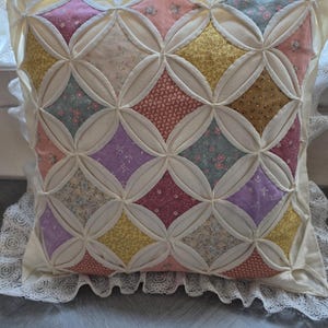 May include: A square throw pillow featuring a cathedral window quilt pattern with cream-colored fabric frames surrounding various floral-printed fabric squares in shades of pink, yellow, purple, and blue, finished with a white lace trim border.
