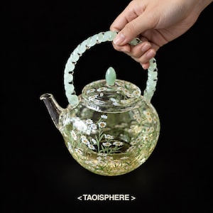 May include: A clear glass teapot featuring a painted pattern of white daisy flowers with green stems and leaves. The teapot has a rounded body, a matching glass lid with a small finial, and a twisted, pale green translucent handle.