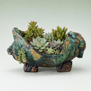 May include: A textured, dark green and brown ceramic planter with four short legs, containing a variety of small green and light purple succulent plants. The planter has a rough, uneven rim and a mottled, multi-colored glaze finish.