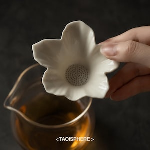 Puede incluir: Un colador de té de cerámica blanca con forma de flor de cinco pétalos y una base perforada para filtrar las hojas de té. El colador está colocado sobre una jarra de vidrio transparente que contiene un líquido de color ámbar. El texto TAOISPHERE es visible.