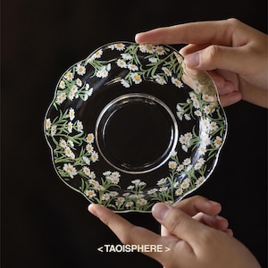 Peut inclure: Une soucoupe en verre transparent à bord festonné, ornée d'un motif répétitif de marguerites blanches à cœur jaune et tiges vertes. Le texte <TAOISPHERE> est imprimé en blanc au centre en bas de l'image sur un fond sombre.