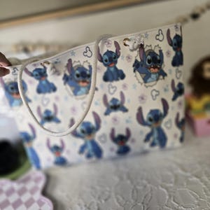 May include: White tote bag featuring a repeating pattern of the blue alien character Stitch from Lilo & Stitch. The bag has a white rope handle and gold-colored grommets. The background is white with small purple flowers and Stitch's face.