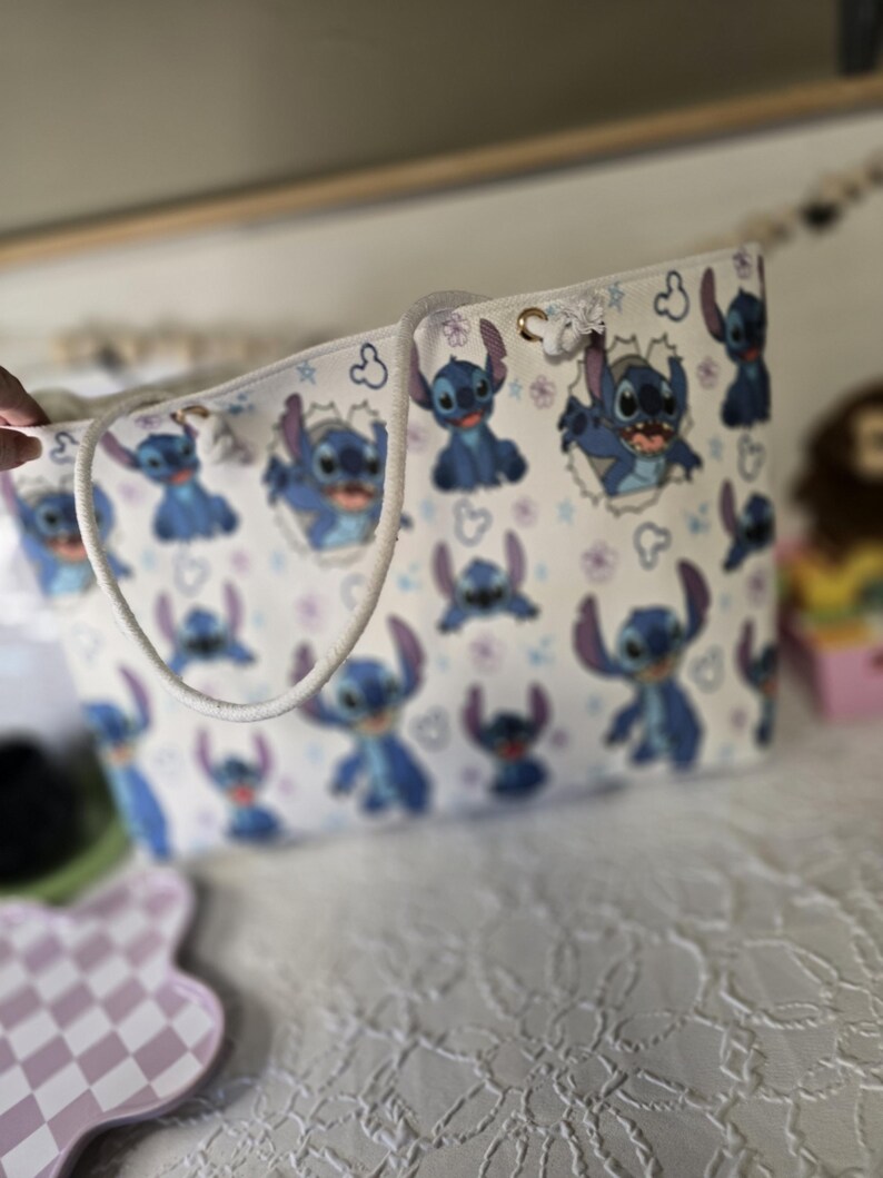 May include: White tote bag featuring a repeating pattern of the blue alien character Stitch from Lilo & Stitch. The bag has a white rope handle and gold-colored grommets. The background is white with small purple flowers and Stitch's face.