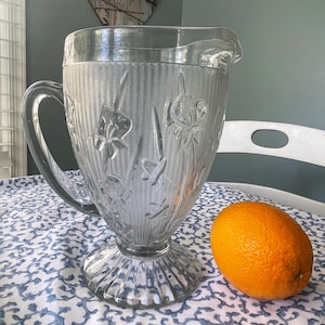 May include: A clear glass pitcher featuring a textured herringbone pattern and embossed floral designs on the exterior. The pitcher has a sturdy handle, a pouring spout, and a wide, fluted pedestal base. An orange is positioned next to the pitcher.