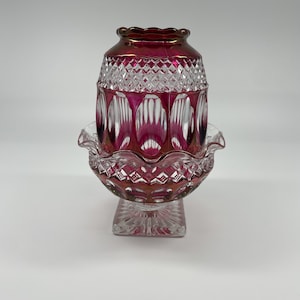 Puede incluir: Un recipiente de dos piezas de cristal tallado en color rojo arándano y transparente, con una sección superior de patrón de diamantes y borde festoneado. La base es un cuenco con borde ondulado, con facetas geométricas y vidrio rojo.