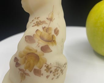 Puede incluir: Una figura de gato de vidrio translúcido de color crema con diseños florales pintados a mano en tonos marrón y bronceado en su espalda. El gato está representado en posición sentada con la cabeza ligeramente girada, mostrando detalles de rosas.