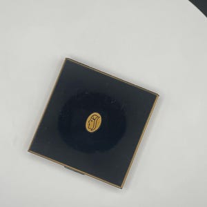 May include: A square black compact case with a gold-toned metal trim. Centered on the lid is an oval gold emblem featuring a line-art profile of a face. The surface of the compact is smooth and dark, contrasting with the metallic gold accents.