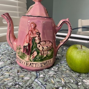 May include: A pink ceramic teapot with a lid, featuring a raised relief scene of a figure and a well with the text REBEKAH AT THE WELL. The teapot has a curved handle and spout with black trim, and is positioned next to a single green apple.