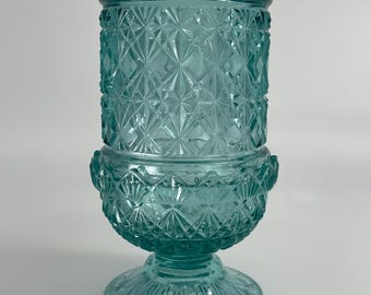 May include: A teal blue glass vessel featuring a two-part construction with a cylindrical top and a rounded base. The surface is decorated with a repeating geometric diamond and starburst pattern, finished with a wide, circular pedestal foot.
