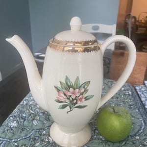 Puede incluir: Una tetera de cerámica de color crema con un motivo floral de flores rosas y hojas verdes en la parte frontal. La tetera tiene una tapa a juego con un borde dorado, un asa curva y una base de pedestal, colocada junto a una manzana verde.
