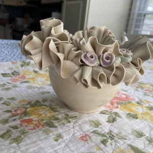 May include: A cream-colored ceramic bowl featuring an elaborate ruffled rim that mimics fabric folds. The exterior of the bowl is smooth, while the top edge is adorned with delicate, hand-sculpted light purple and pale green floral accents.