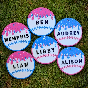 May include: Six circular ornaments featuring a white baseball design with red stitching. Each ornament has a colorful icing drip pattern with sprinkles in pink or blue. The names Ben, Memphis, Audrey, Libby, Liam, and Alison are printed in black.