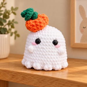May include: A white crocheted ghost plush toy with a rounded body and small arms. It features two black plastic eyes, pink stitched cheeks, and a textured orange pumpkin with green leaves attached to the top of its head.