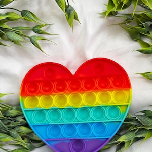 May include: A heart-shaped fidget toy in a rainbow of colors, including red, orange, yellow, green, blue, and purple. The toy has a series of circular bubbles that can be pushed in and out. The background includes green leafy sprigs.