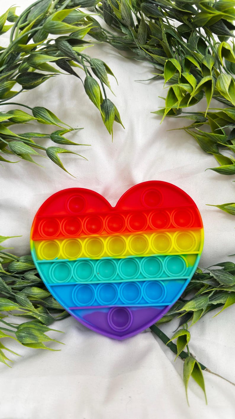 May include: A heart-shaped fidget toy in a rainbow of colors, including red, orange, yellow, green, blue, and purple. The toy has a series of circular bubbles that can be pushed in and out. The background includes green leafy sprigs.